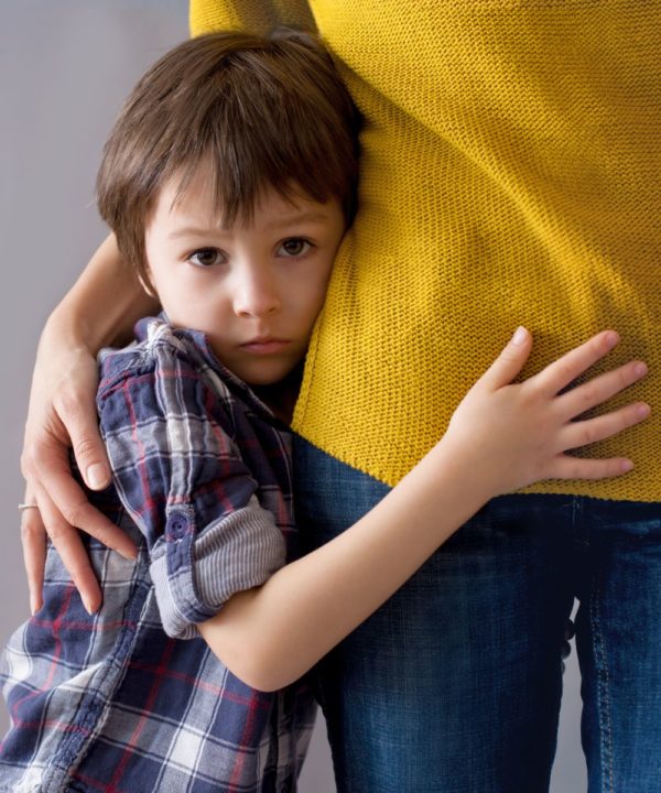 a child that looks worried wearing a flannel shirt, as he clings to his mom's hip